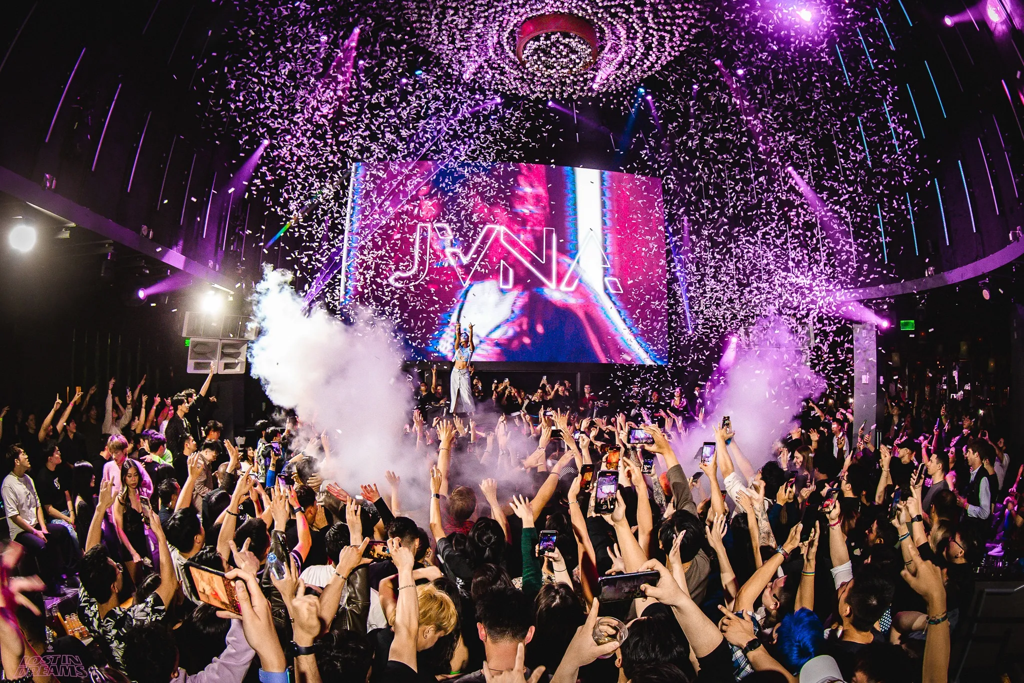 A large crowd at a concert or nightclub in Costa Mesa dances and cheers with hands raised, surrounded by purple lights, confetti, and smoke. A performer is on stage in front of a big screen displaying the name JVNA—perfect for a Costa Mesa travel blog highlight.
