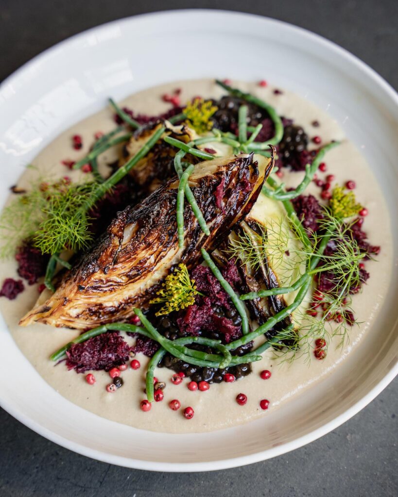 A white bowl with grilled endive on a creamy puree, topped with sea beans, dill, lentils, pink peppercorns, and beet relish—an artful dish perfect for a Costa Mesa travel blog, beautifully arranged on a dark surface.