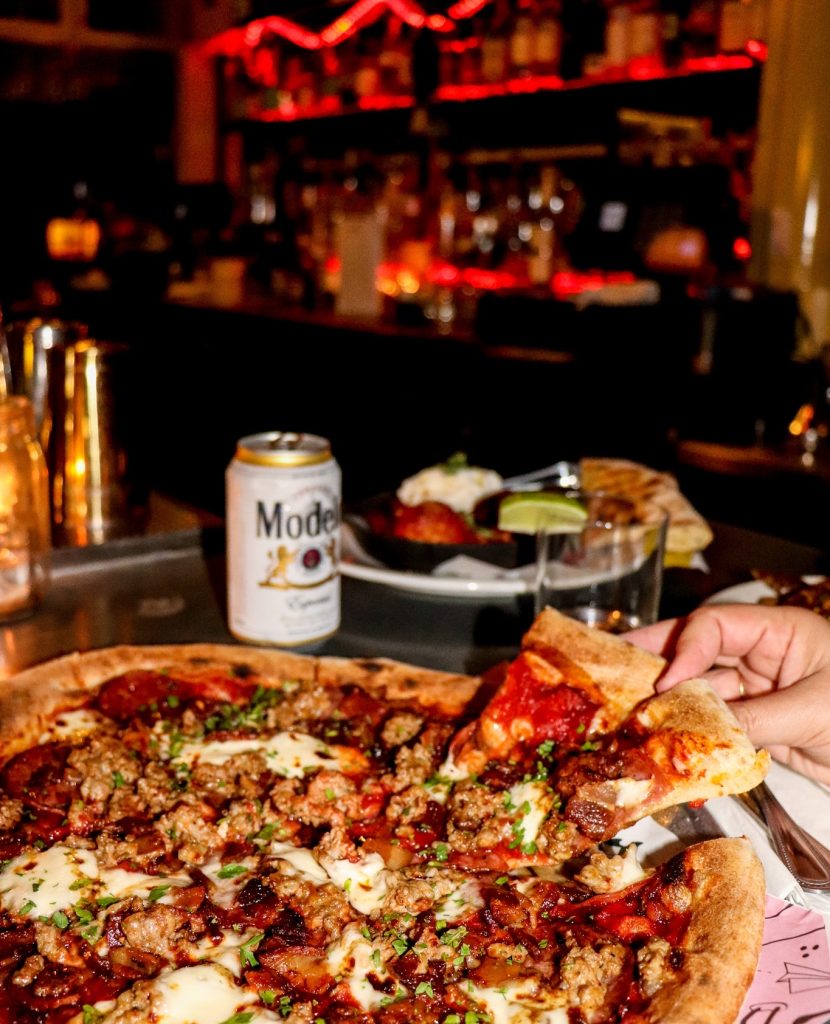 A hand lifts a slice of meat-topped pizza from a tray in a dimly lit bar with red lights. A can of Modelo beer and a plate of food are on the table in the background.