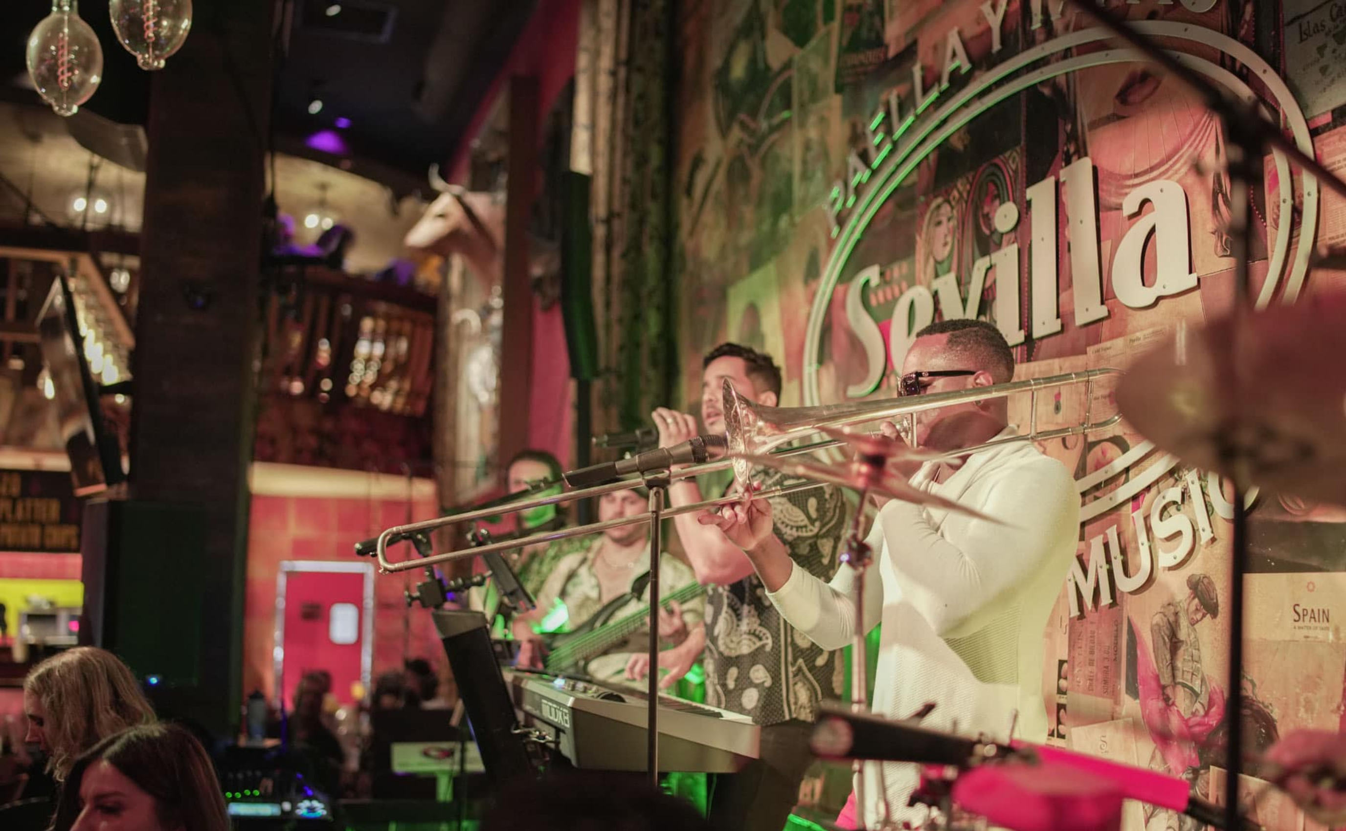 A band performs live on stage at a vibrant venue, with a trombone player in the foreground and colorful lights illuminating the scene. A wall in the background displays the word Sevilla.