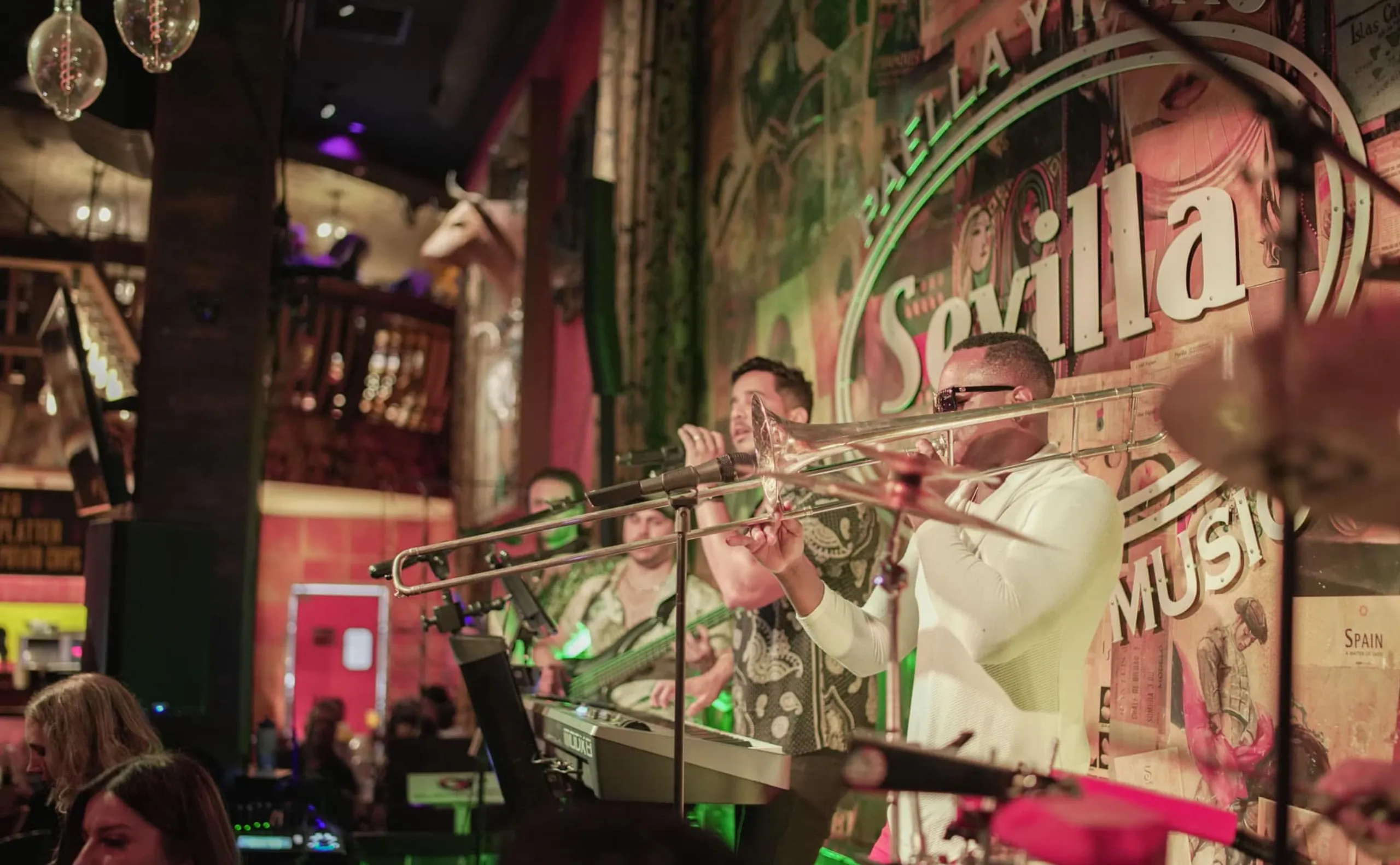A band performs live on stage at a vibrant venue, with a trombone player in the foreground and colorful lights illuminating the scene. A wall in the background displays the word Sevilla.