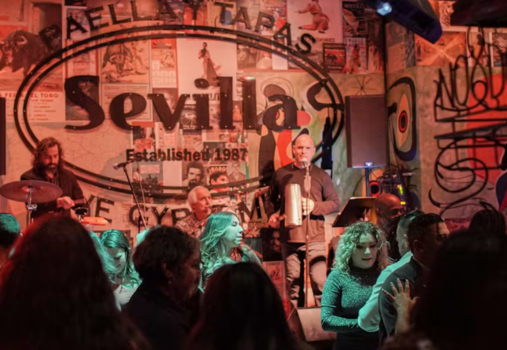 A live band performs on stage at Sevilla, a Spanish-themed restaurant and bar, while people dance and mingle under warm lighting. The walls are decorated with posters and vibrant artwork.