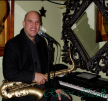 A smiling man in a black suit sits at a keyboard with a saxophone, holding a microphone. There is an ornate mirror and decorations in the background. The name Robert Martell is faintly visible in the corner.