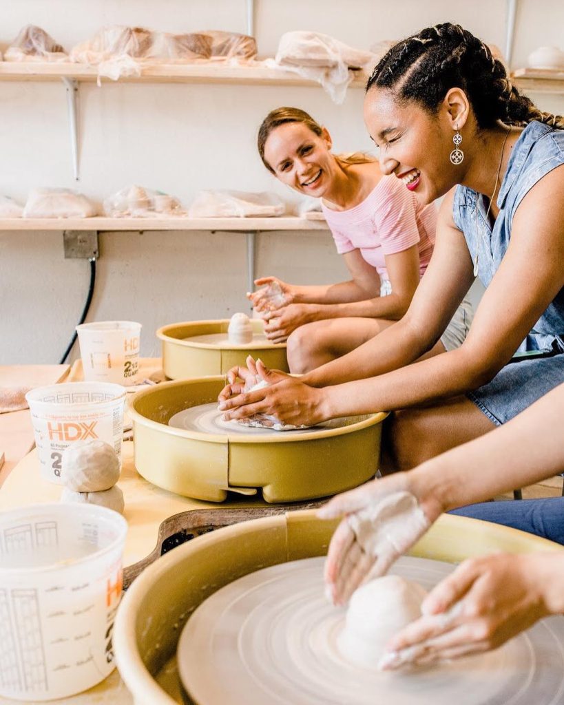 Three women smile and laugh while shaping clay on pottery wheels in a bright, cheerful studio. Shelves with pottery and plastic containers are in the background.