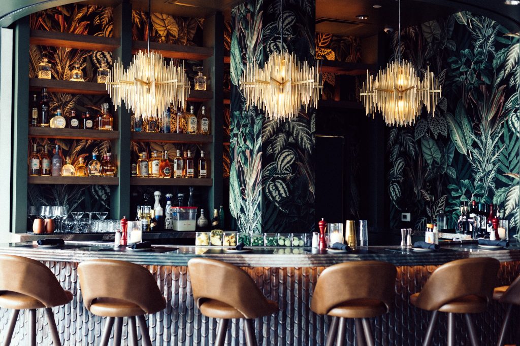 A stylish bar with brown leather stools, bottles and glasses on wooden shelves, and two ornate chandeliers. The background features dark, leafy wallpaper, giving the space a modern and tropical feel.