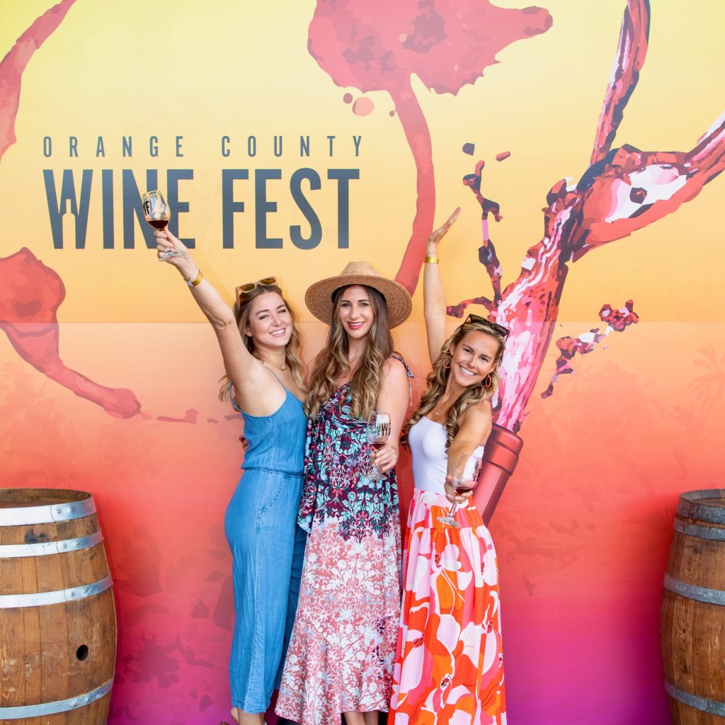 Three women in colorful summer outfits smile and hold up wine glasses in front of an Orange County Wine Fest backdrop, with wine barrel props on each side and splashes of wine illustrated on the background.