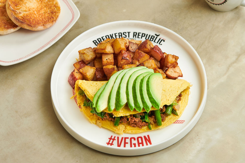 A vegan omelet topped with avocado slices, filled with vegetables and plant-based meat, served with diced, roasted potatoes on a white plate labeled Breakfast Republic #Vegan in Costa Mesa. An English muffin is on a separate plate nearby.