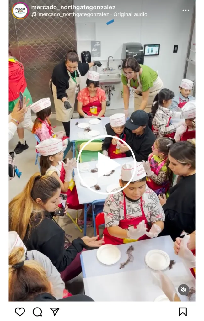 Children wearing chef hats and aprons sit and stand around a table making food, supervised by adults in a kitchen setting. Some kids shape ingredients with their hands, while others watch or wait with bowls and utensils.