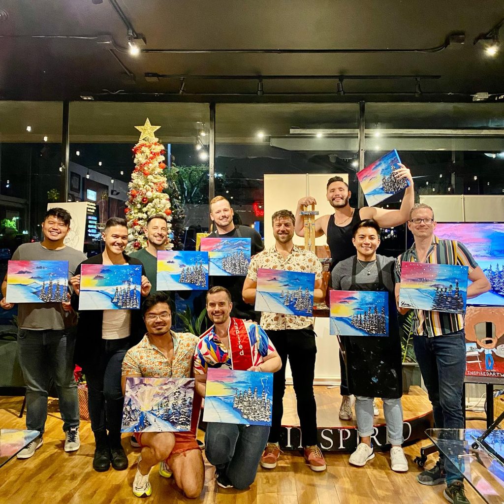A group of twelve people pose indoors with their completed paintings, each featuring a colorful sky and trees. A decorated Christmas tree is in the background, and everyone appears cheerful and proud of their artwork.