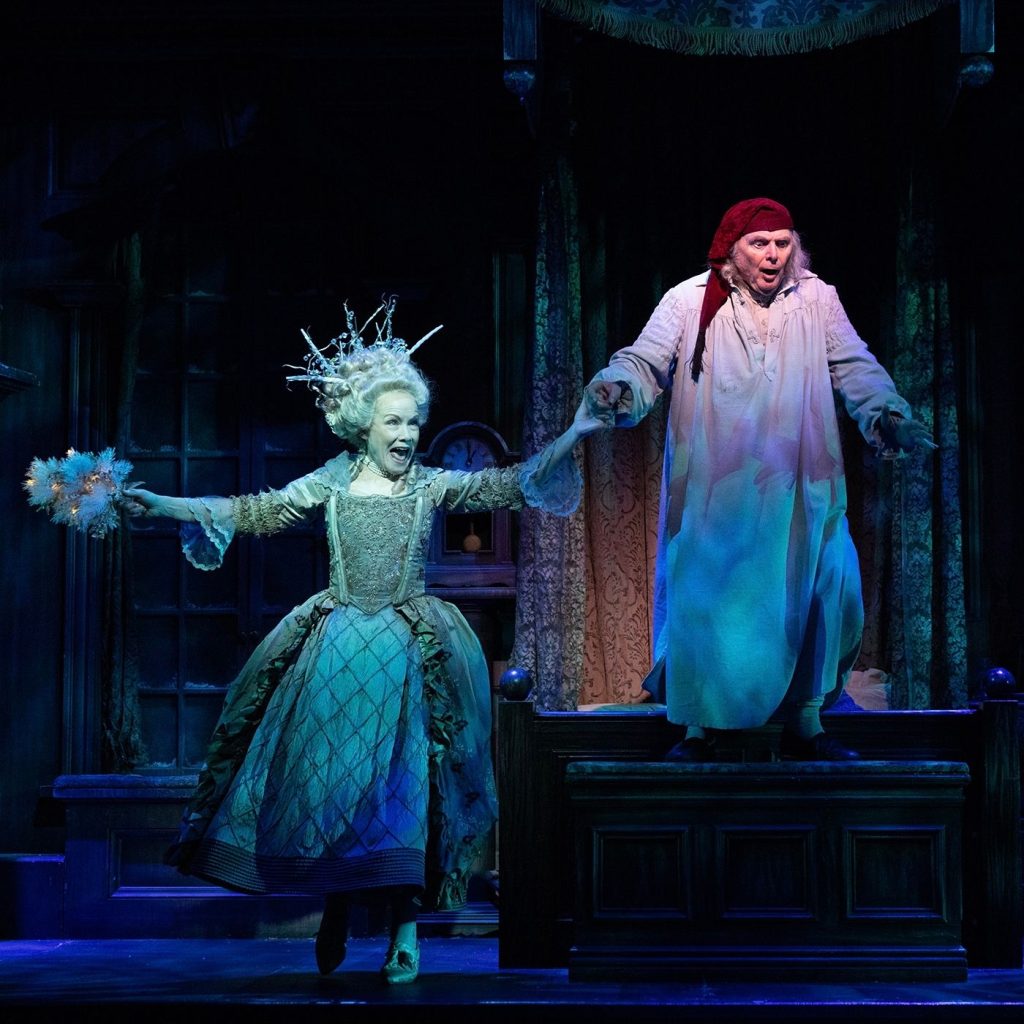 Two actors perform on stage; one wears a lavish, icy blue gown and crown, while the other, dressed in a nightgown and nightcap, stands on a bed looking surprised. The scene is dramatically lit with blue highlights.