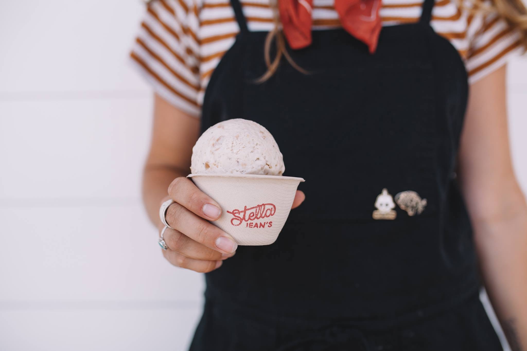 A person in a striped shirt, red scarf, and black apron holds a cup of Stella Jean’s ice cream—Costa Mesa’s favorite—showing only their torso and hand against a white background.
