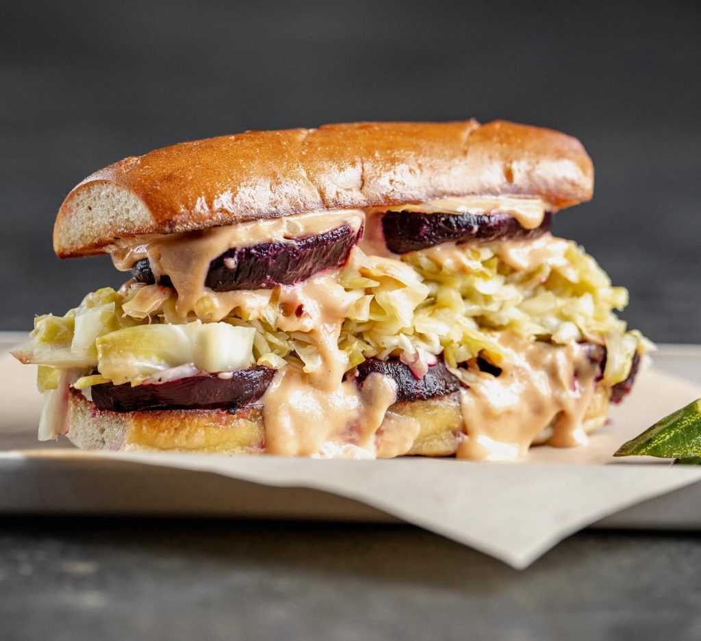 A close-up of a sandwich filled with sliced beets, shredded cabbage, and creamy sauce, served on a glossy, toasted bun, placed on a piece of parchment paper with a dark background.