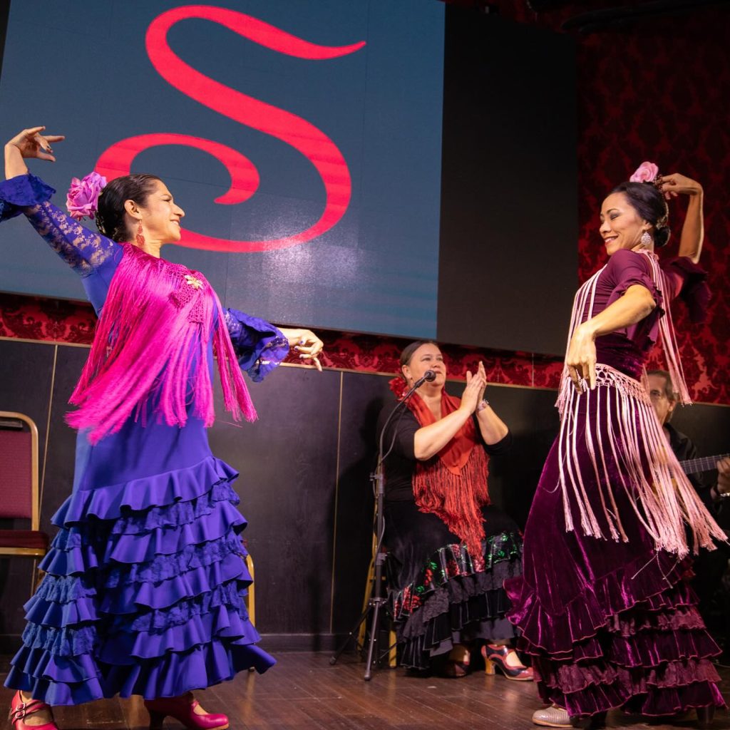 Two women in colorful ruffled dresses and shawls perform a Flamenco dance on stage, while a seated singer claps along. A bold red S is displayed on a screen behind them.