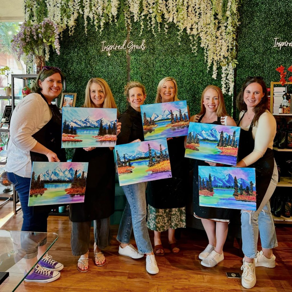 Six smiling women stand indoors holding colorful landscape paintings they created, featuring mountains, trees, and lakes. The background has a green plant wall with white flowers and the words Inspired Goals.