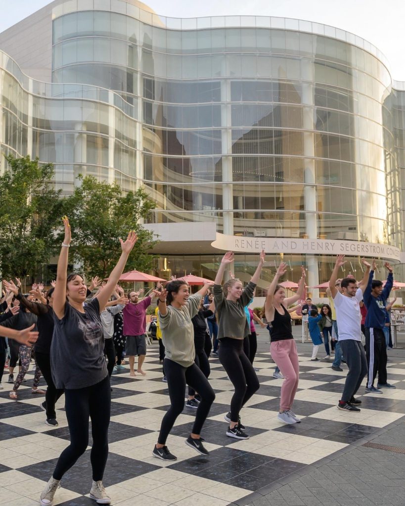 A group of people dance energetically with raised arms on a checkered outdoor plaza in front of a modern glass building, with others watching and trees nearby.