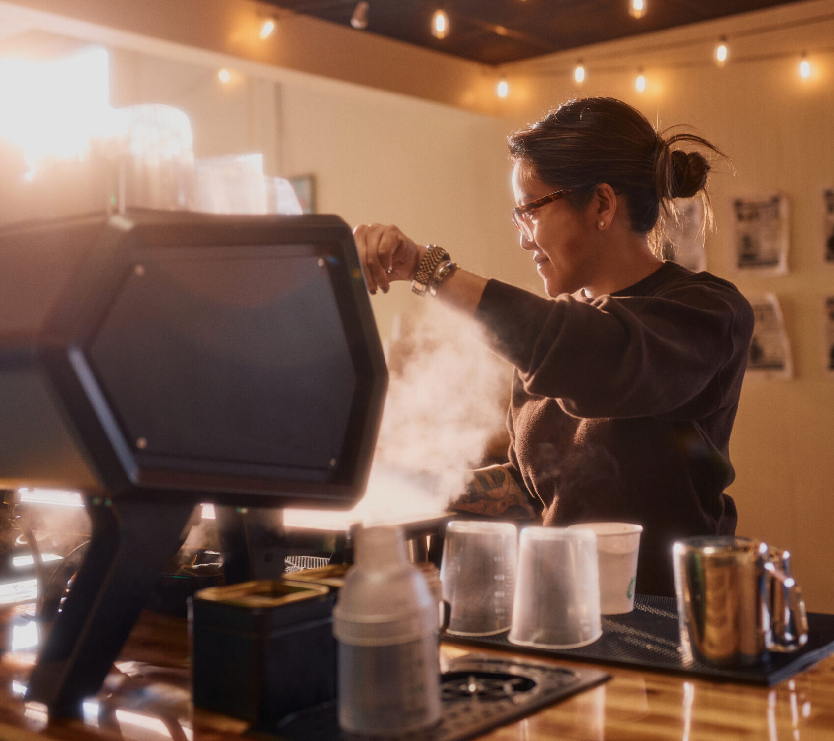 A barista with glasses and tied-back hair works behind a coffee machine, surrounded by mugs and steam, in a warmly lit Costa Mesa café adorned with string lights and a cozy atmosphere.