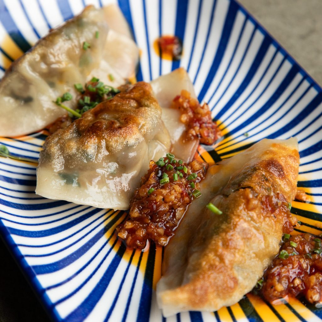 Three pan-fried dumplings with golden, crispy edges are served on a blue and white striped plate, garnished with chopped herbs and accompanied by a chunky red chili sauce.