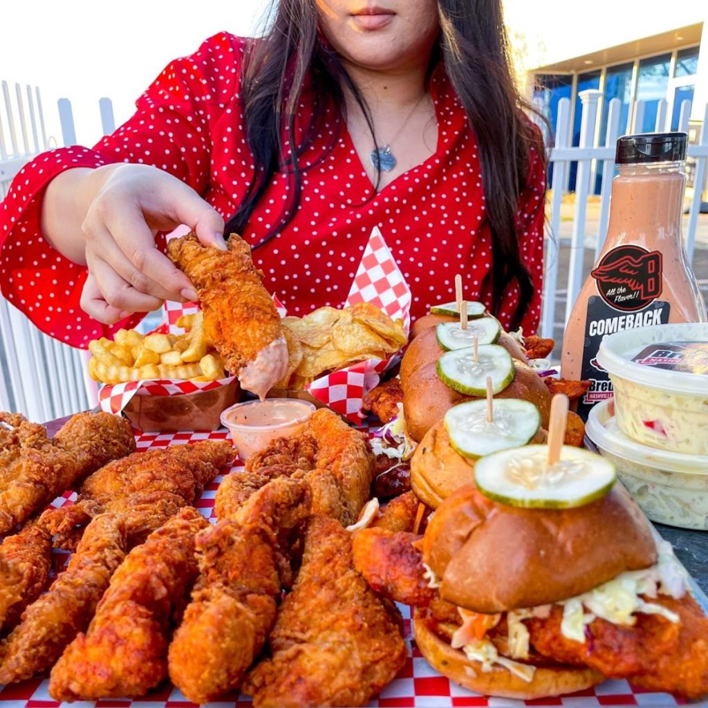 A woman in a red polka dot shirt holds a fried chicken tender over a table filled with fried chicken, crinkle fries, sliders topped with pickles, coleslaw, and a bottle of comeback sauce.