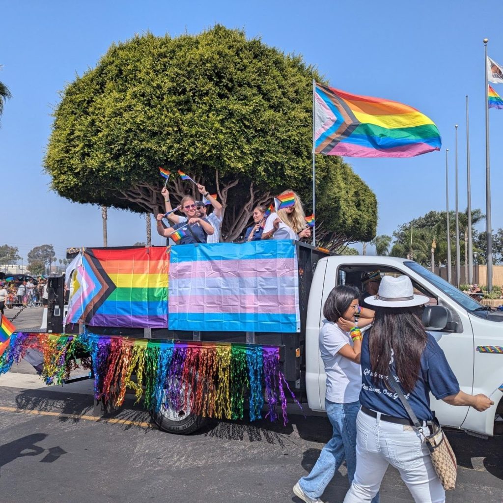 A white truck decorated with rainbow, transgender, and progress pride flags and colorful streamers carries people waving pride flags during a parade. Two women walk beside the truck; a large tree is in the background.