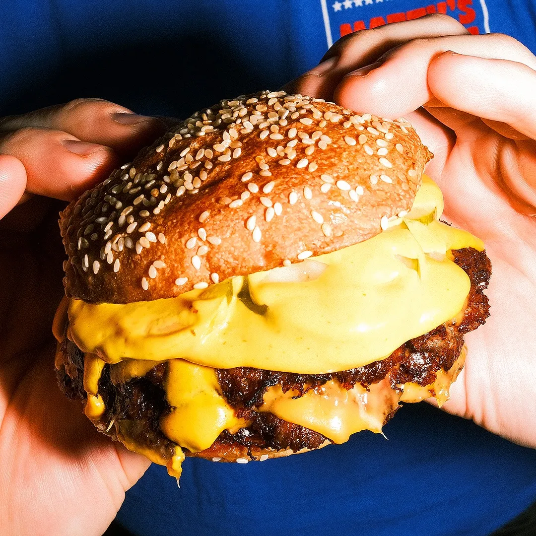 Close-up of hands holding a large cheeseburger with a sesame seed bun, two beef patties, and melted yellow cheese dripping down the sides. The background is blue.