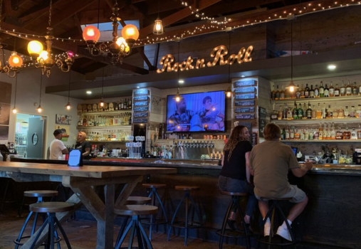 A cozy bar with string lights and colorful hanging lamps, a sign reading Rock n Roll, a TV above the bar, and people sitting on stools chatting. Bottles line the shelves behind the bar.