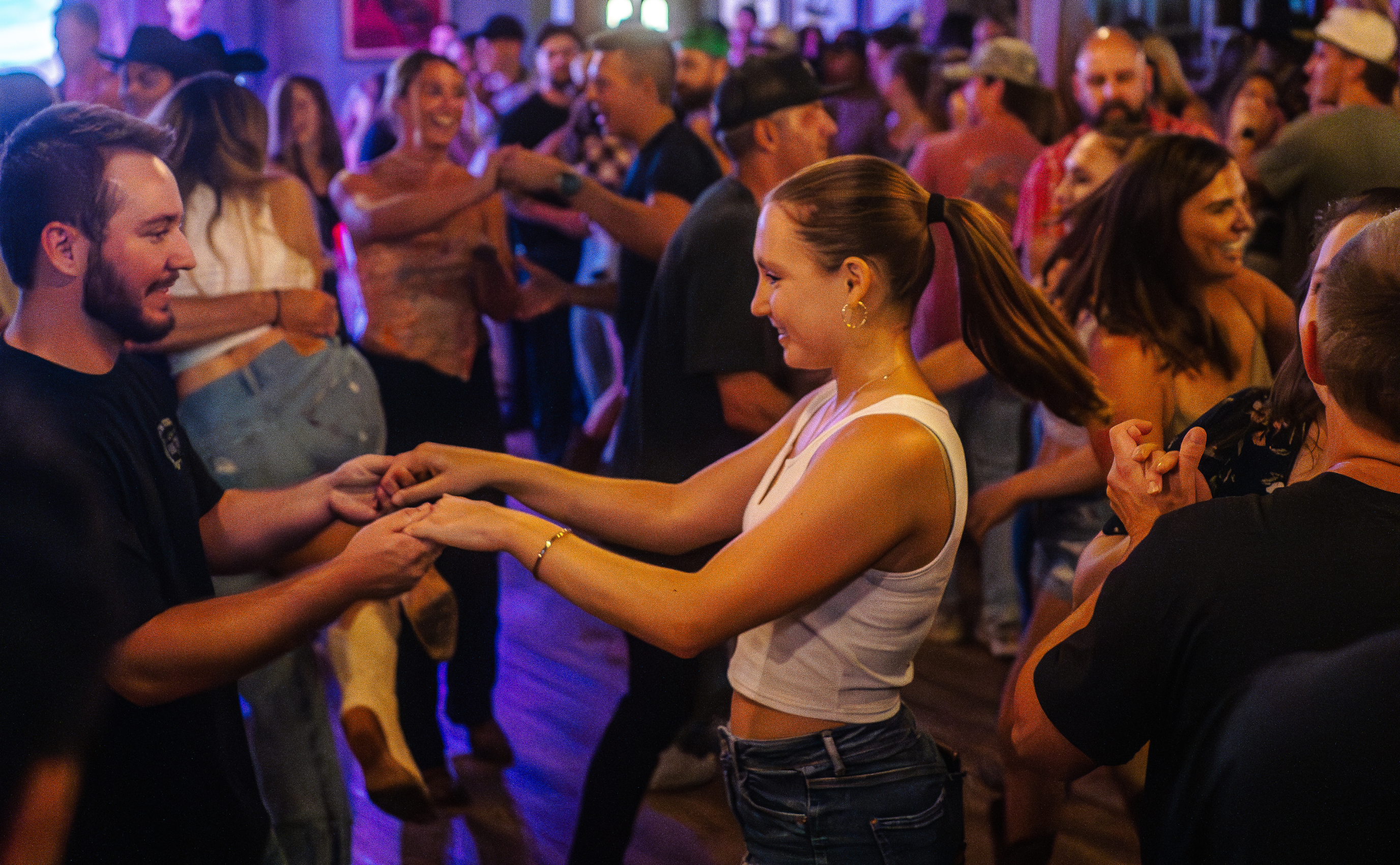 A couple dances closely in a dimly lit bar, surrounded by other people. The man in the foreground wears a black t-shirt, and the woman wears a white tank top and jeans.