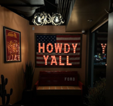 A dimly lit room with an American flag on the wall and illuminated letters spelling “HOWDY Y’ALL.” A red bench labeled “FORD” sits below, and a framed cowboy picture and cactus are visible.