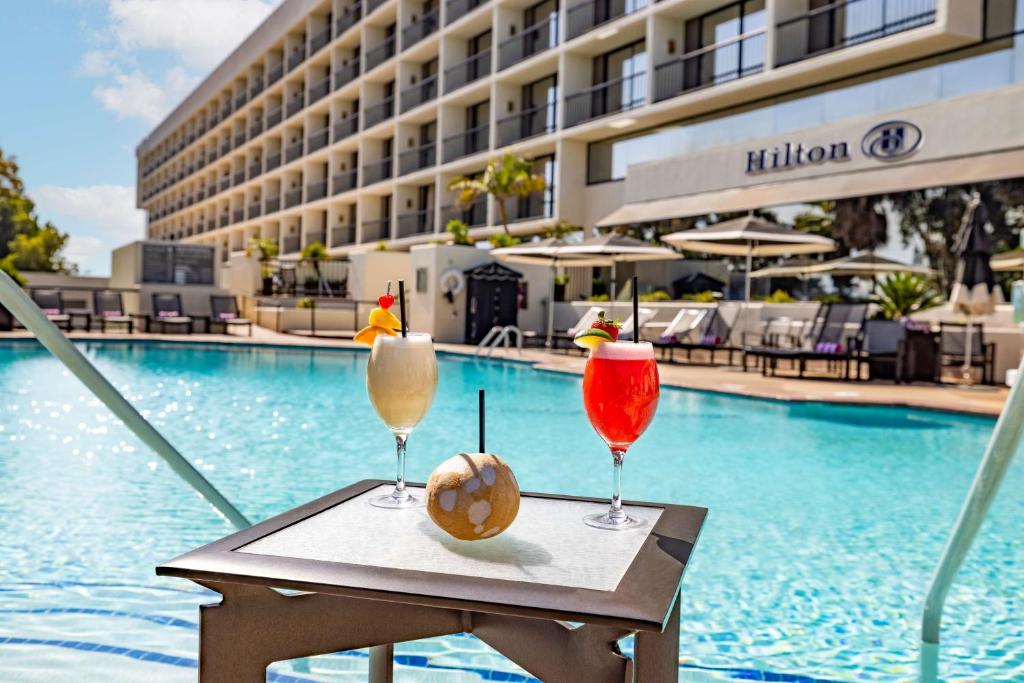 Two colorful cocktails and a coconut drink on a small table by a sunny outdoor pool with lounge chairs, umbrellas, and the Hilton hotel building in the background.