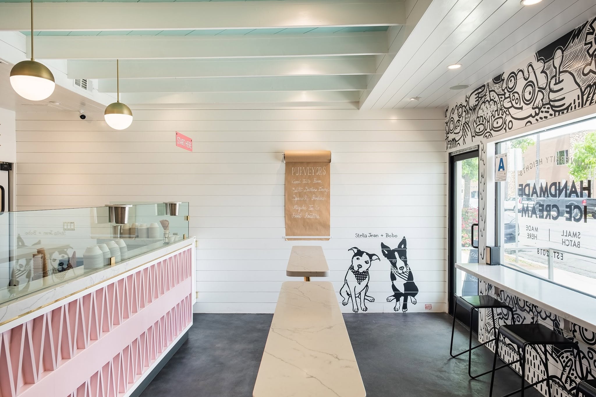 Bright Stella Jeans Ice Cream Costa Mesa shop with white shiplap walls, marble tables, pink counter, and a whimsical dog and cat mural. Large windows and pendant lights create an airy, inviting atmosphere.