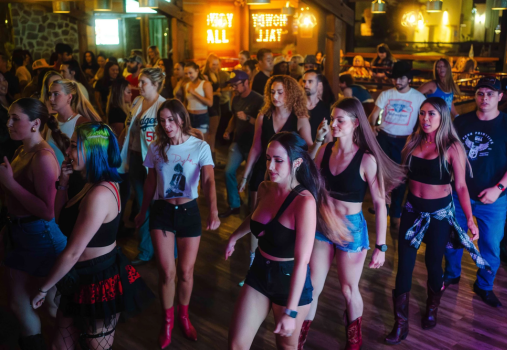 A group of people, mostly women, line dancing indoors at a lively bar or club with neon signs and wooden decor, dressed in casual and country-style outfits.
