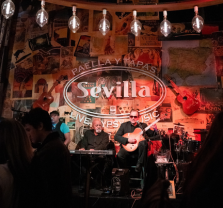 Two musicians perform on a small stage at Sevilla, a live music and tapas venue, with guitars and posters decorating the wall behind them. The audience watches in a dimly lit, cozy atmosphere.