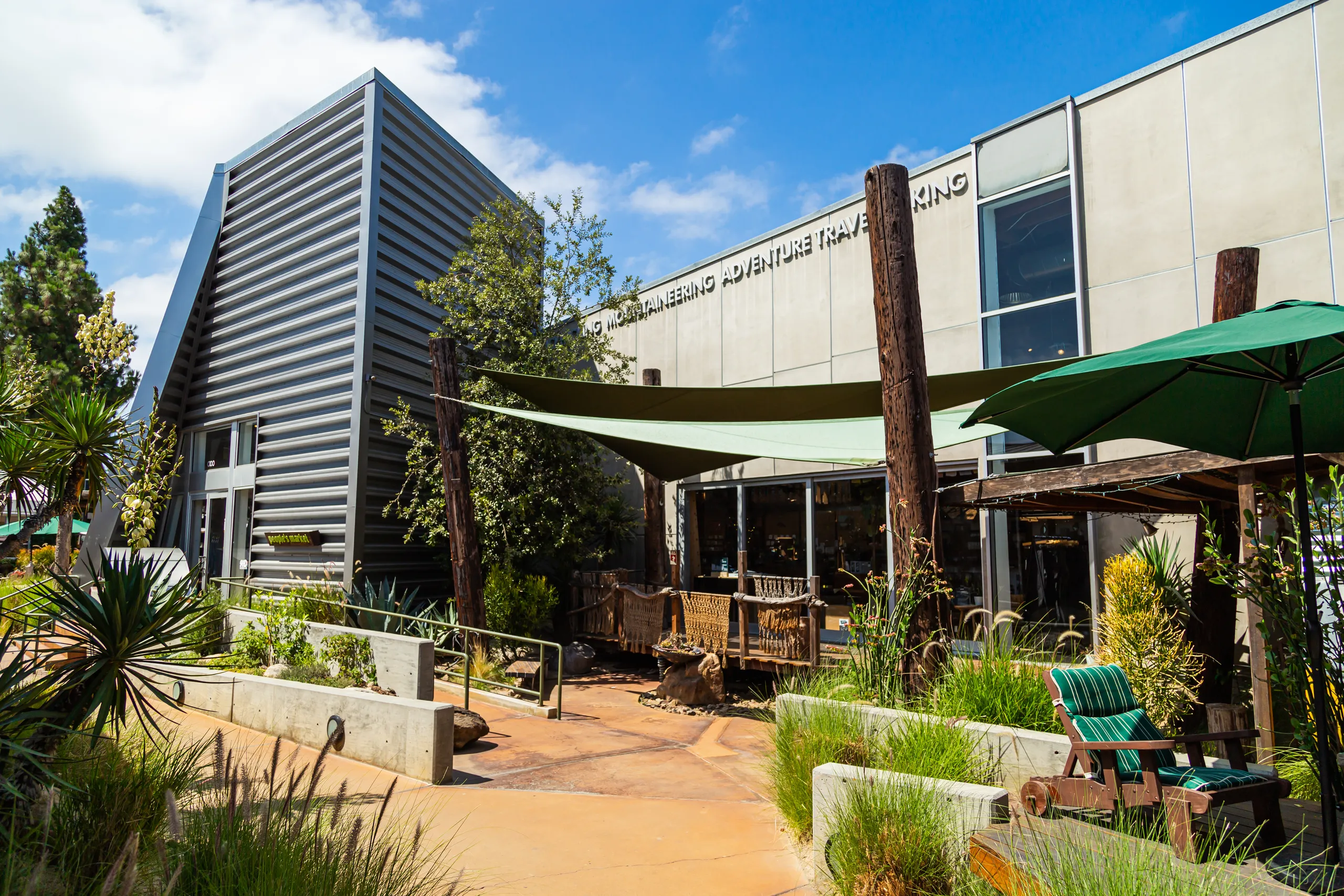 A modern building with large windows, metal siding, and outdoor seating areas surrounded by greenery under a sunny blue sky.