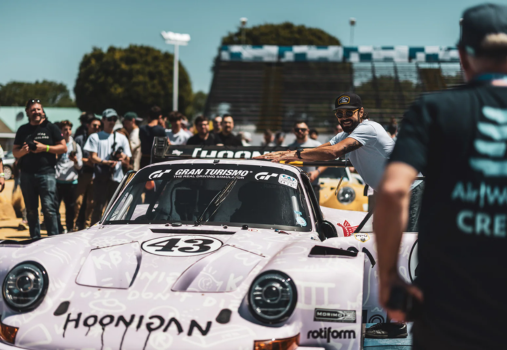 A crowd surrounds a pink race car with the number 43, covered in graffiti-style writing. A smiling man leans on the cars roof, while another person in a CREW shirt stands in the foreground. Spectators watch from bleachers.