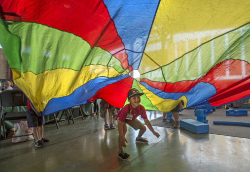 A young child crouches and smiles beneath a colorful parachute held up by other children indoors, with light streaming through large windows in the background.