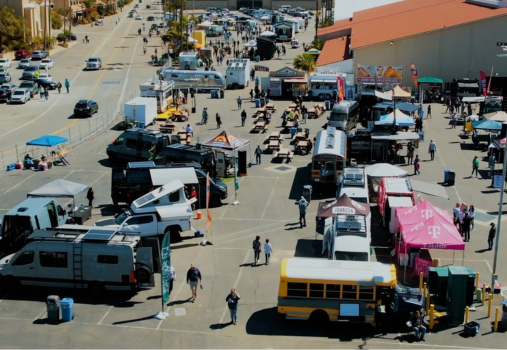 A busy outdoor event with various camper vans, tents, vendor booths, and picnic tables. People walk around and gather near vehicles and exhibits in a large parking lot on a sunny day.