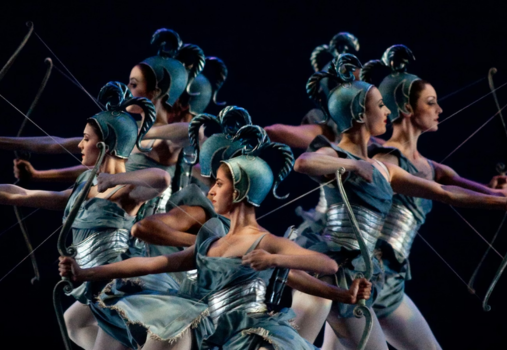 A group of ballet dancers in metallic blue costumes and feathered helmets pose as archers, drawing invisible bows in unison against a dark background.
