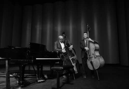 Two musicians in suits stand onstage beside a grand piano, each playing a double bass. The background is softly lit with vertical panels, and the scene is in black and white.