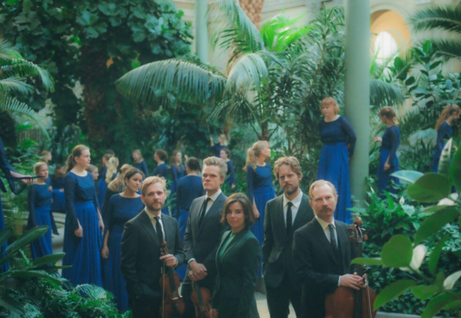 A group of five musicians holding string instruments stand among lush green plants, while several women in long blue dresses are seen walking and standing in the background inside a conservatory.