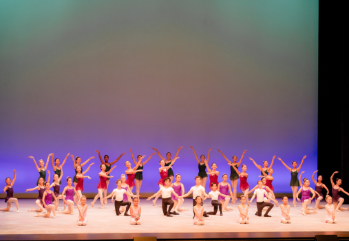 A large group of ballet dancers in colorful costumes pose on stage, arms raised gracefully, against a gradient blue and green backdrop during a performance.