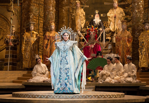 A woman in ornate, pale blue traditional costume stands on stage before a group of performers in gold and white robes and elaborate headdresses, set against a lavish, golden, throne-like backdrop.