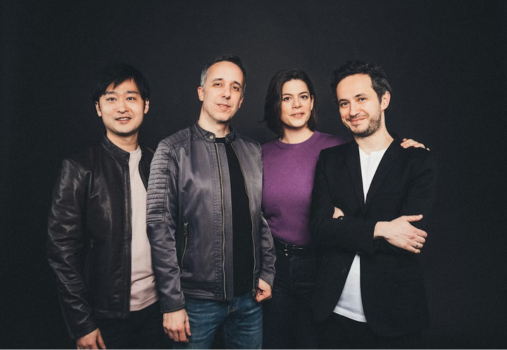 Four people stand together against a dark background. Three men and one woman, all smiling, wear casual jackets and shirts. The woman, in a purple top, stands between two of the men.