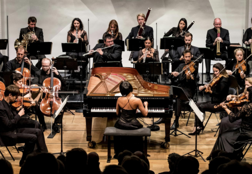 A pianist in a black dress performs on a grand piano with a symphony orchestra, including string, woodwind, and brass musicians, all dressed in black, on a concert hall stage.