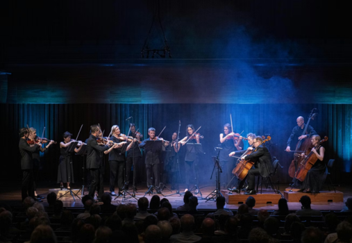 A classical string orchestra performs on stage under blue lighting, with violinists, cellists, and a double bassist playing. The audience is seated in the foreground, and mist adds atmosphere to the scene.