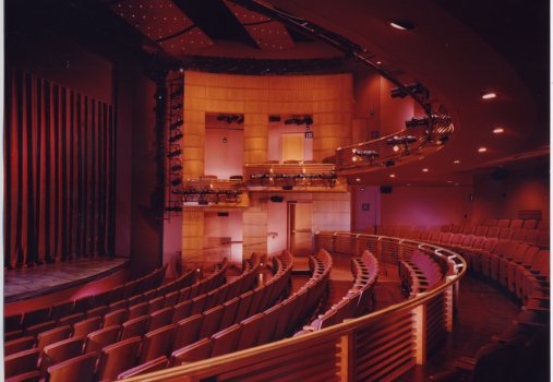 A modern theater interior with curved rows of empty red seats, illuminated balconies, and a stage with closed curtains on the left under warm, ambient lighting.