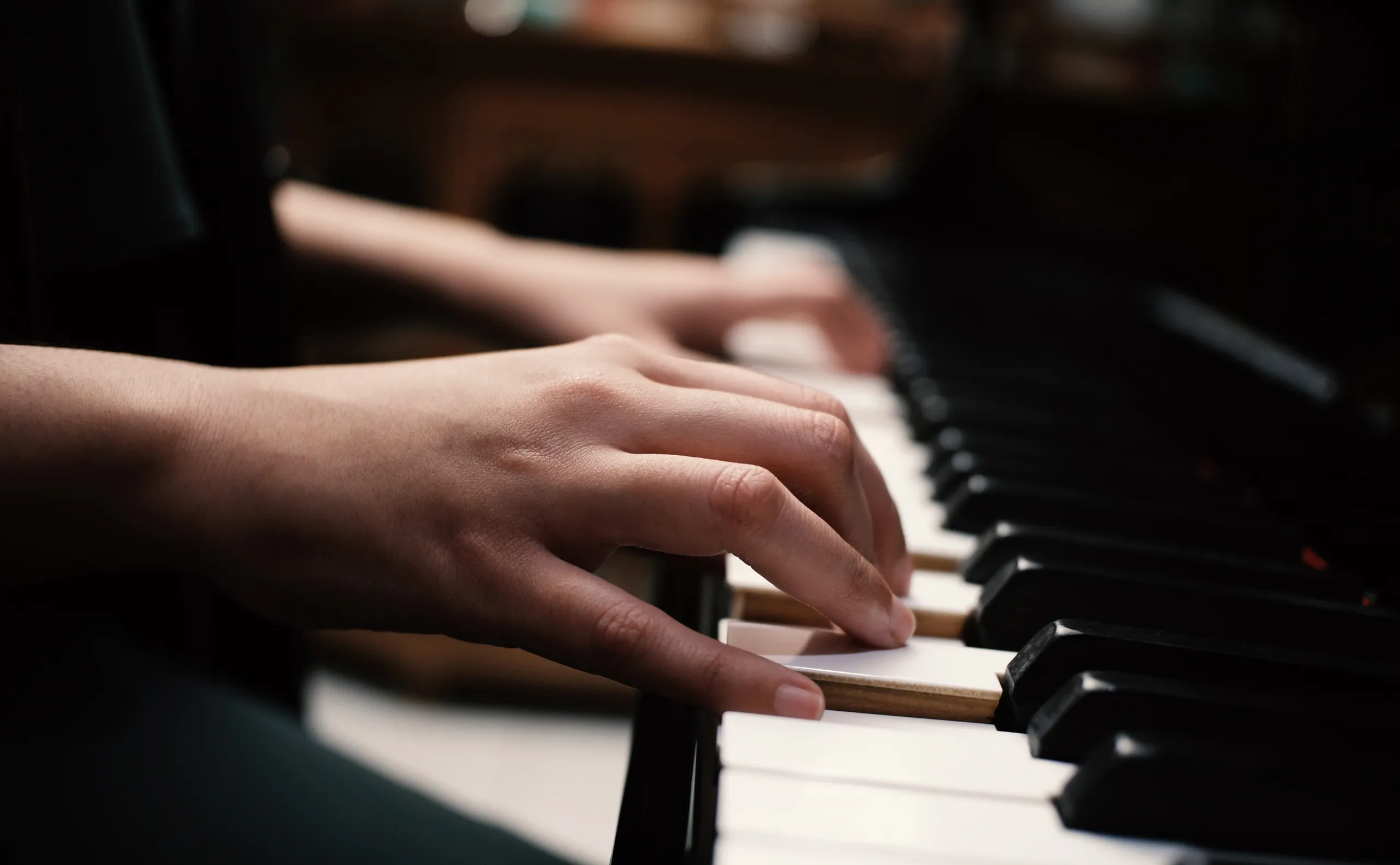 Close-up of a persons hands playing a piano, with fingers pressing down on the keys. The background is softly blurred, drawing focus to the piano and hands.