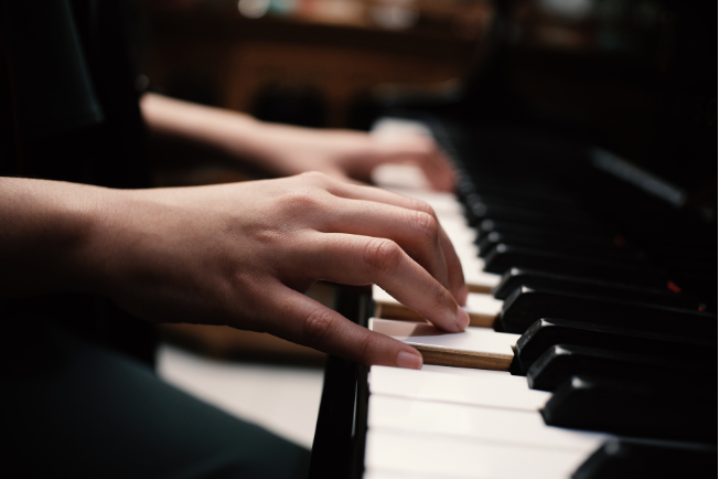 Close-up of a persons hands playing the keys of a piano, with fingers pressing down on the black and white keys. The background is softly blurred, emphasizing the focus on the hands and the piano keyboard.