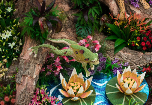 A detailed frog sculpture made from plants and flowers appears to leap over a pond with two large floral water lilies, surrounded by vibrant foliage and colorful blossoms.