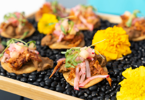 Small tostadas topped with shredded meat, pickled onions, and microgreens are arranged on a tray covered with black beans and decorated with bright yellow marigold flowers.
