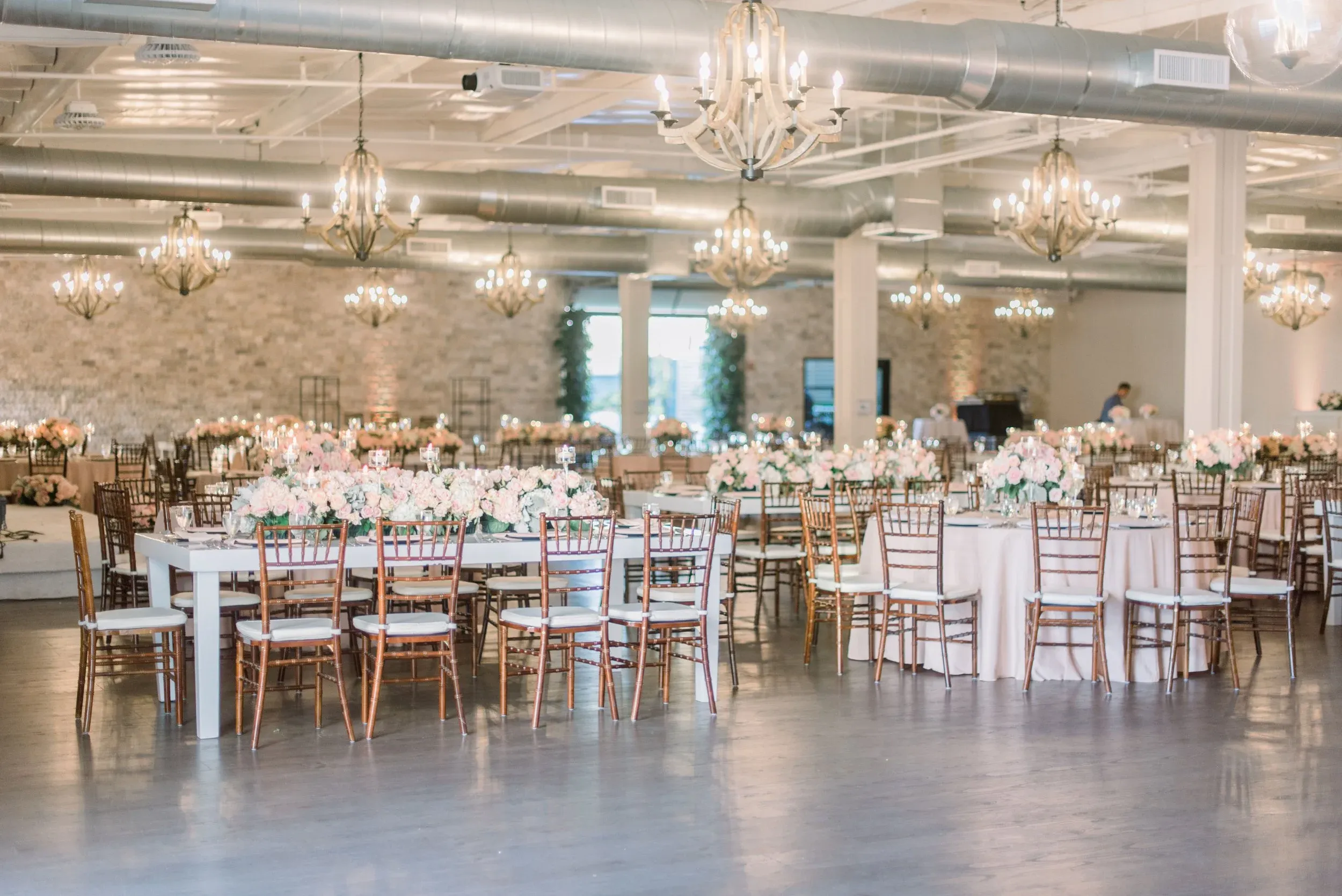 Elegant banquet hall decorated for an event, featuring round and rectangular tables with floral centerpieces, wooden chairs, chandeliers, and soft lighting in a spacious, modern setting—perfect for those seeking top wedding venues Costa Mesa offers.