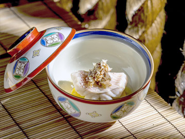 A decorative bowl with a partially open lid contains a delicately plated piece of fish topped with garnish, placed on a bamboo mat with warm light streaming in—a dish inspired by Costa Mesa, City of the Arts.